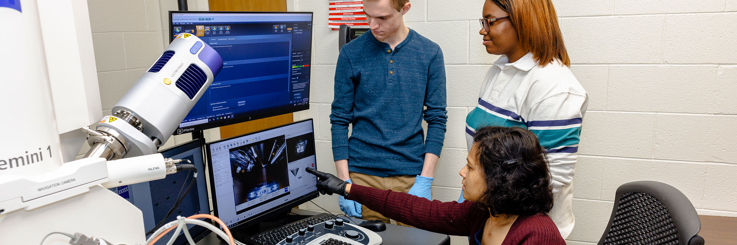 Dr. Mahua Biswas and two students in a lab, interacting with computer monitors and scientific equipment.